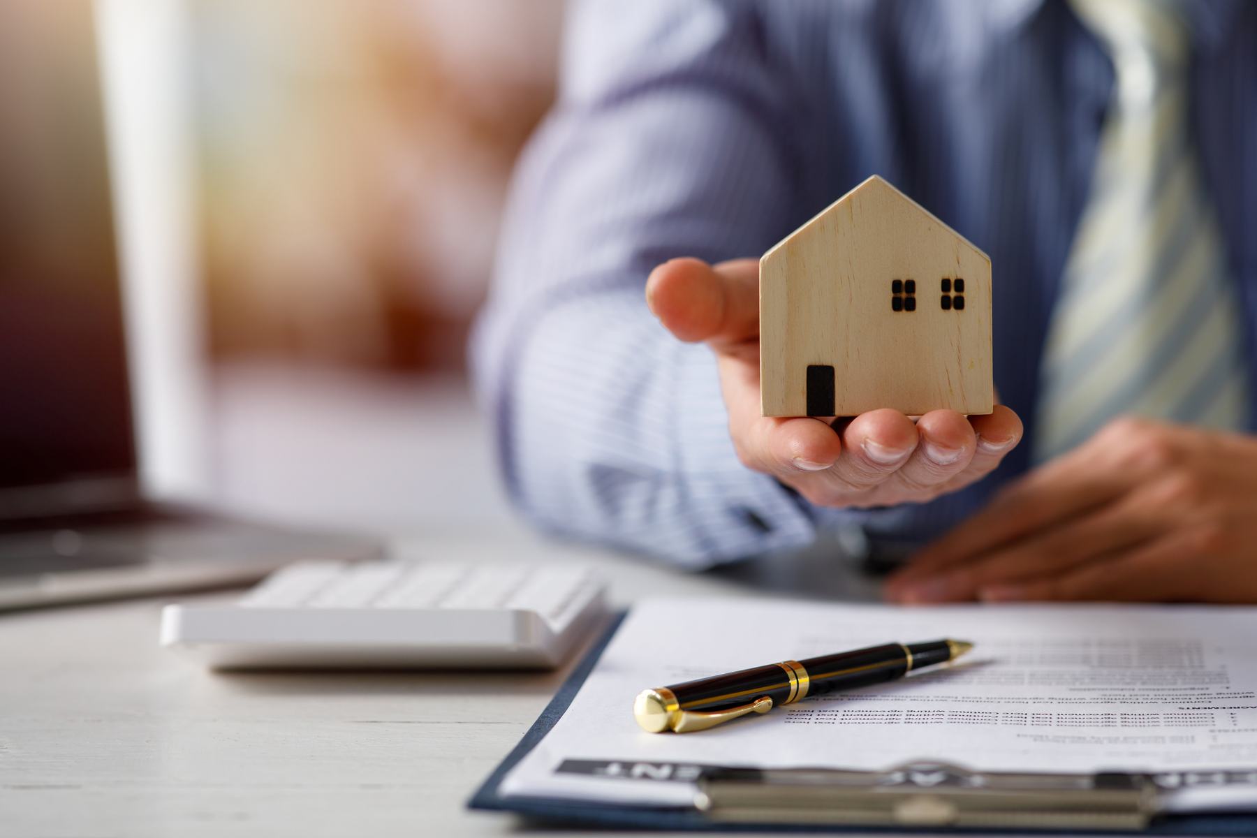 A person holding a small wooden model house above a signed agreement and pen on a desk.